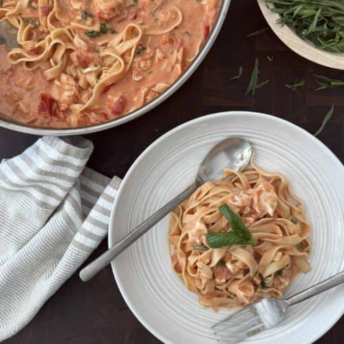 Lobster pasta is plated on a white plate with utensils and the large skillet of more lobster pasta is behind.