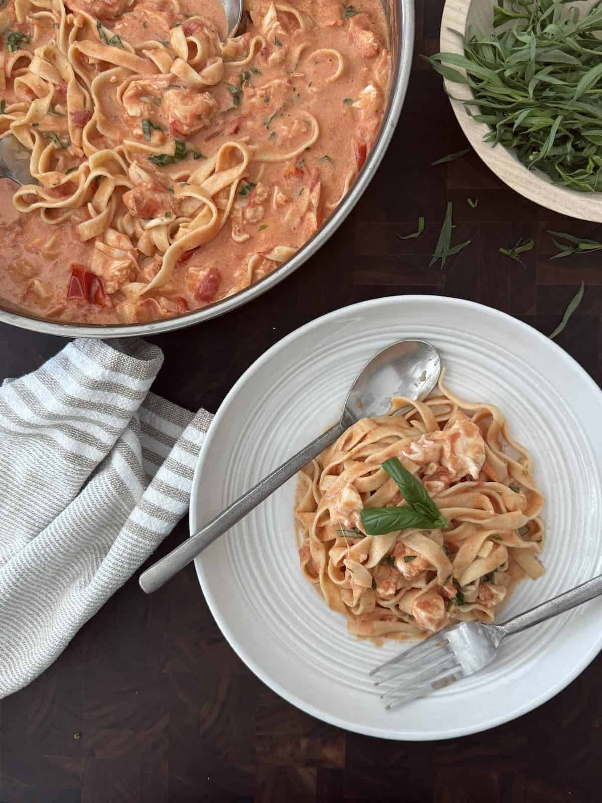 Lobster pasta is plated on a white plate with utensils and the large skillet of more lobster pasta is behind.