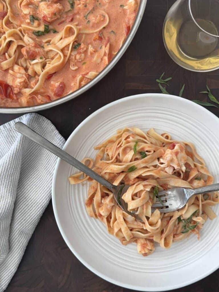 Lobster pasta is on a white plate with utensils. The large skillet of more pasta and a glass of white wine is behind.
