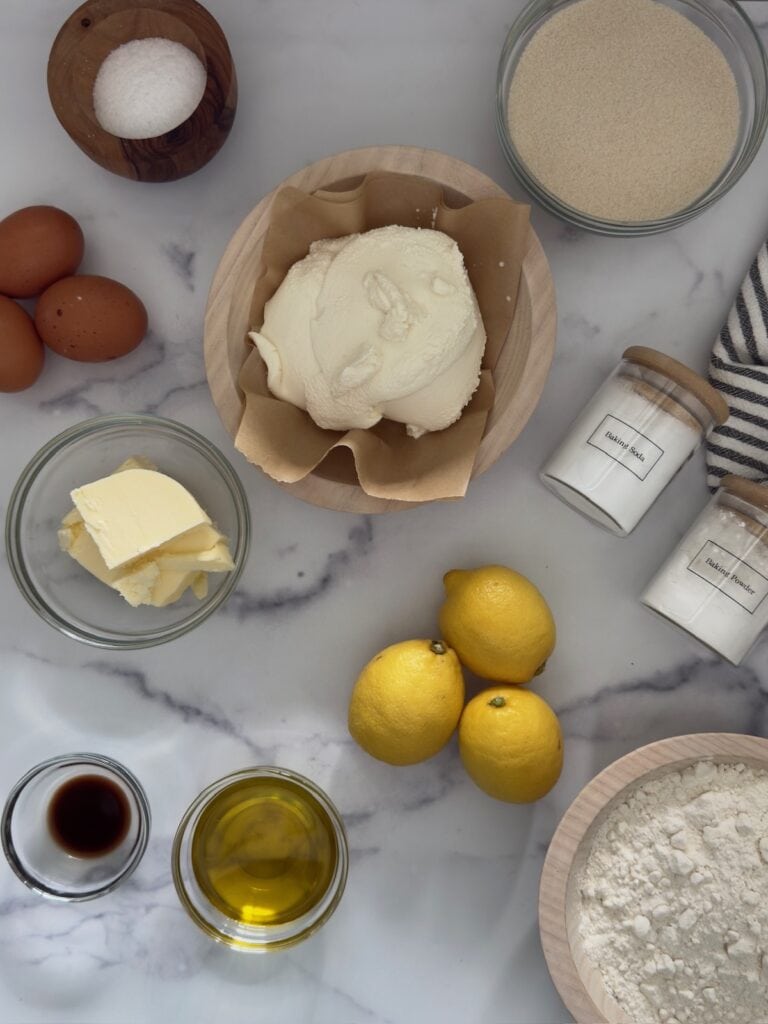 Ingredients for lemon ricotta cake in glass and wood bowls. The ingredients from top to bottom include salt, flour, ricotta, baking soda, baking powder, eggs, ricotta, butter, lemon, olive oil, flour, and vanilla extract.