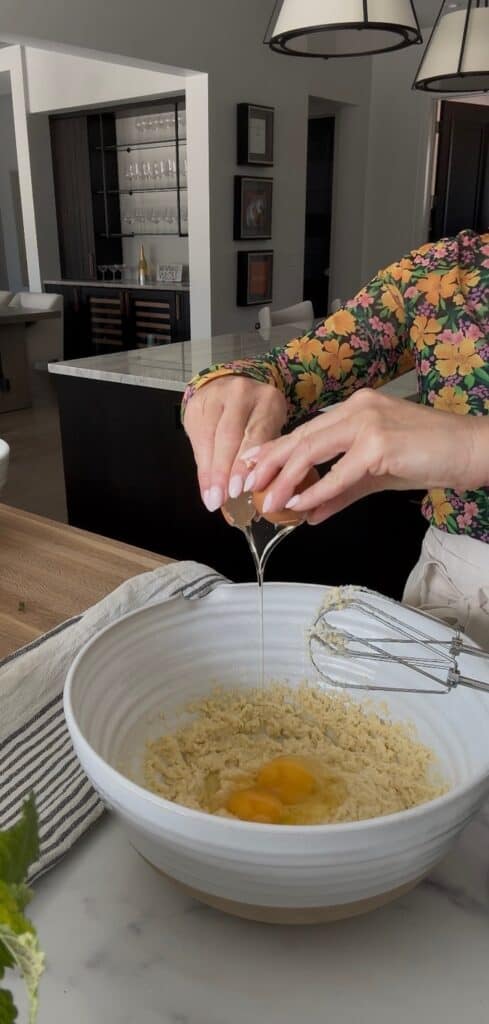 Two hands are cracking eggs into a large white mixing bowl with the lemon ricotta cake batter.