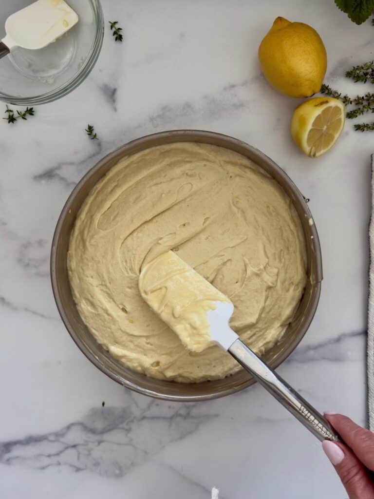 Lemon ricotta cake batter is being smoothed by a white spatula into the springform cake pan. A hand is holding the spatula.