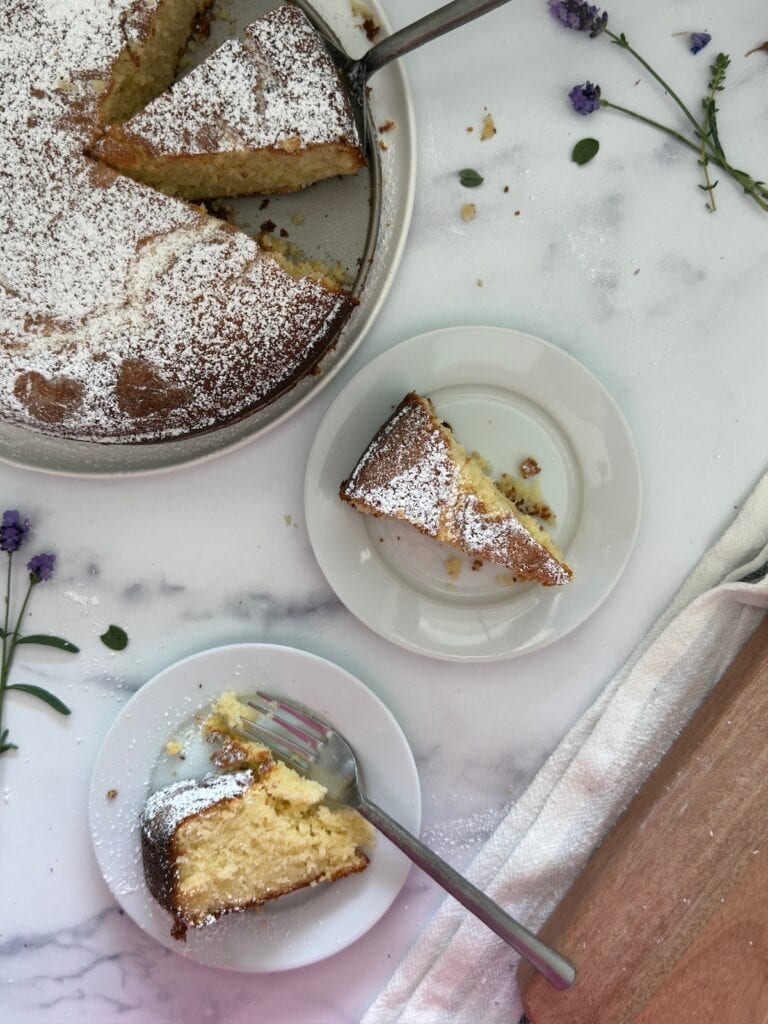 Two slices of lemon ricotta cake are on white plates. The whole lemon ricotta cake is in the background on a white plate.