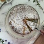 Finished lemon ricotta cake is sliced and on a plate with a cake serve holding one piece. A cut lemon is in the background.