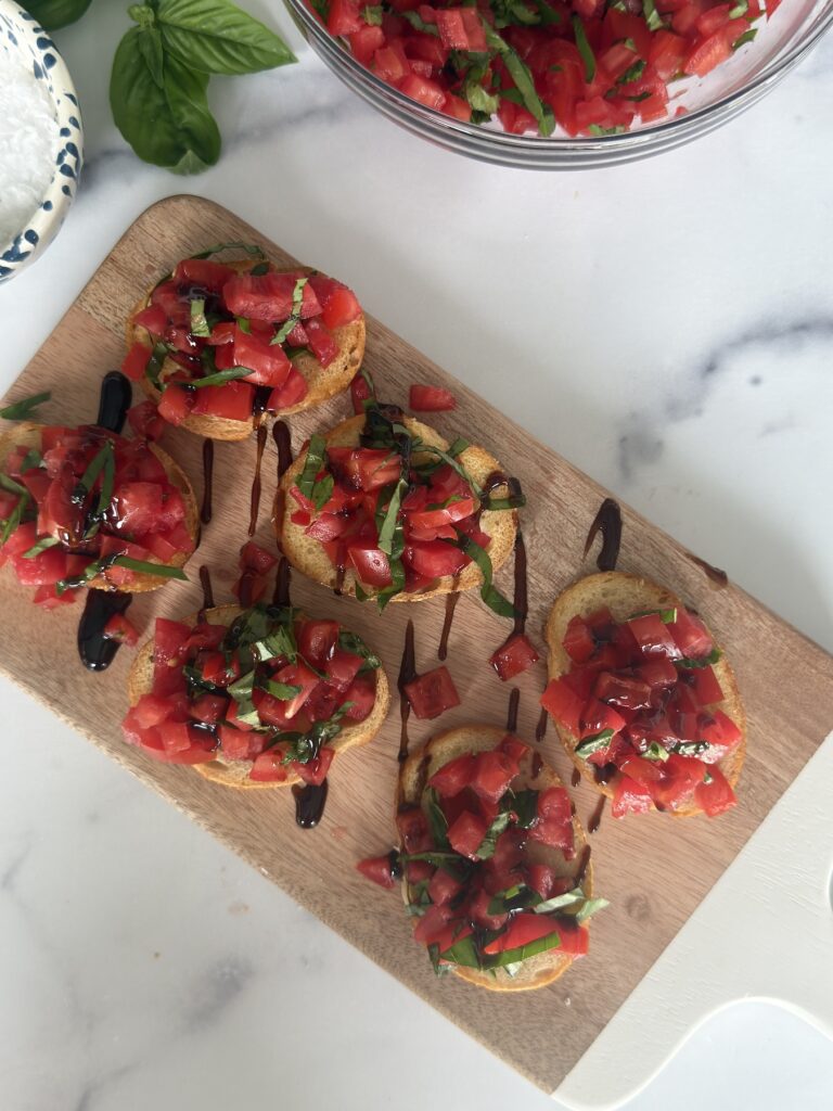 Overhead view of Bruschetta al Pomodoro with balsamic glaze on a cutting board, surrounded by fresh basil and tomato topping.