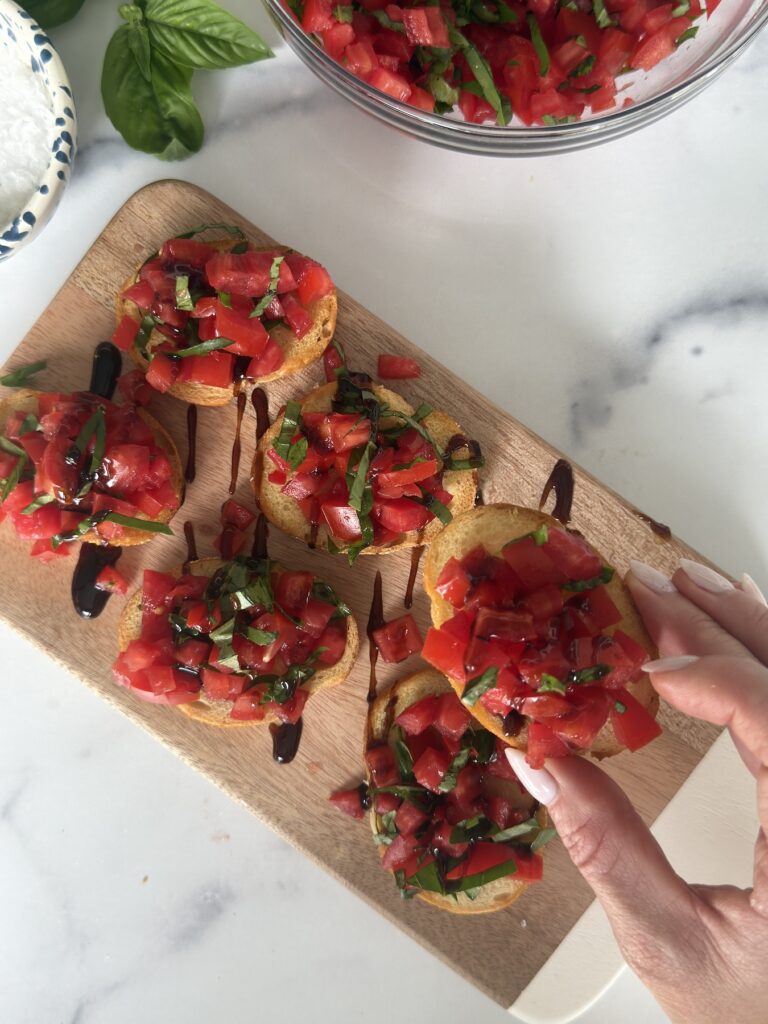 Hand reaching for a slice of Bruschetta al Pomodoro topped with tomatoes, basil, and balsamic glaze.