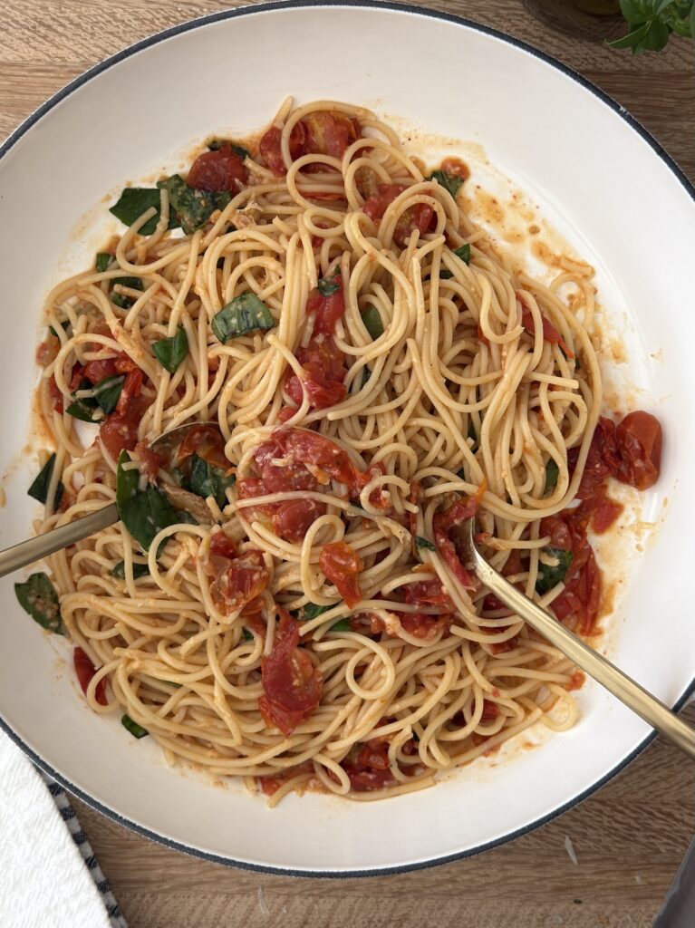 Spaghetti with roasted tomatoes and fresh basil in a white bowl, part of a simple burrata pasta recipe served on a wooden table.