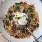 Overhead shot of burrata pasta with roasted tomatoes, topped with burrata cheese, basil, and grated Parmesan, served in a white bowl on a wooden table.