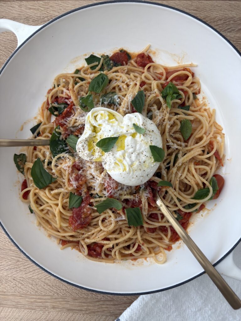 Overhead shot of burrata pasta with roasted tomatoes, topped with burrata cheese, basil, and grated Parmesan, served in a white bowl on a wooden table.