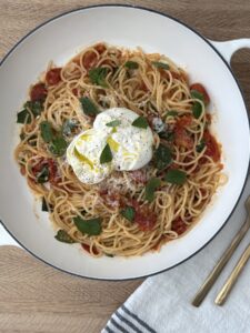 Overhead view of burrata pasta with roasted tomatoes, fresh basil, and Parmesan served in a white bowl with gold utensils on a linen napkin.