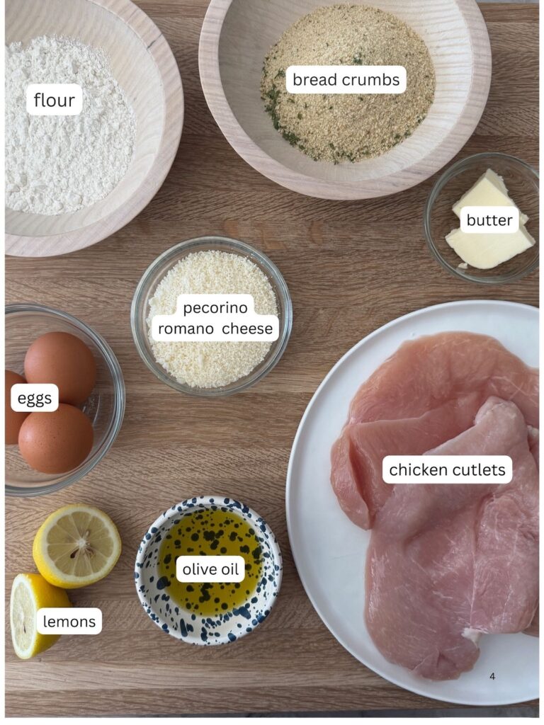 Ingredients for Italian chicken cutlets, including raw chicken breasts, breadcrumbs, flour, eggs, Parmesan cheese, lemon, parsley, and butter arranged on a wooden surface.