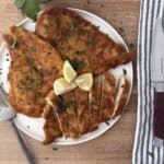 Sliced Italian chicken cutlet on a white plate, garnished with parsley and lemon wedges, served with a striped napkin and fork.
