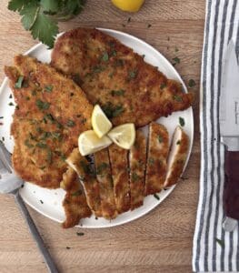 Sliced Italian chicken cutlet on a white plate, garnished with parsley and lemon wedges, served with a striped napkin and fork.