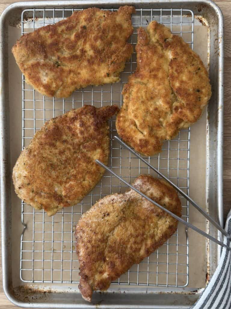 Freshly fried chicken cutlets are resting on a wire rack over a baking sheet to drain excess oil.