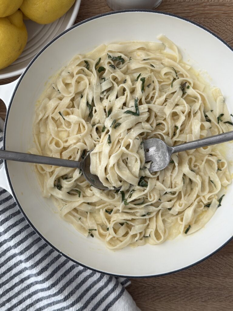 Creamy lemon spaghetti served in a white bowl with fresh herbs, with a fork and knife twirling the pasta, and a bowl of lemons in the background.