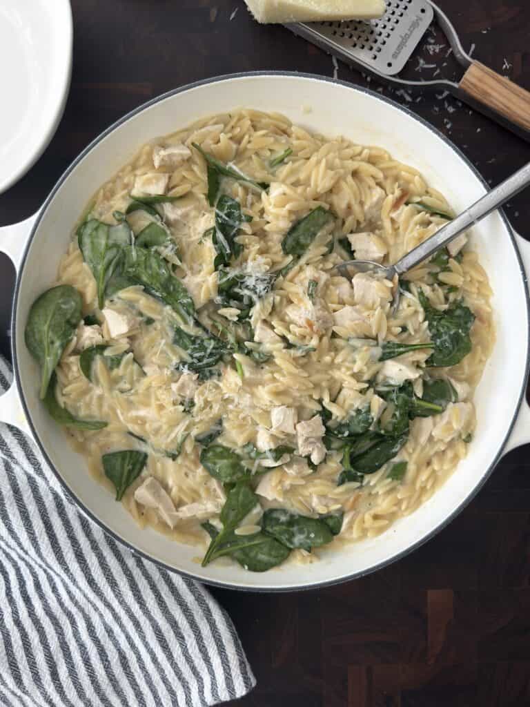 Overhead photo of the Lemon Chicken orzo in a white skillet with a Parmesan cheese grater and a linen napkin on a dark wood cutting board.