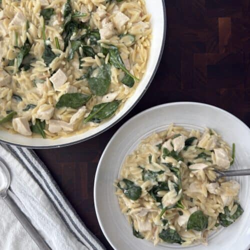 Overhead photo of the Lemon Chicken orzo in a white skillet and a serving bowl with a linen napkin and serving spoons.