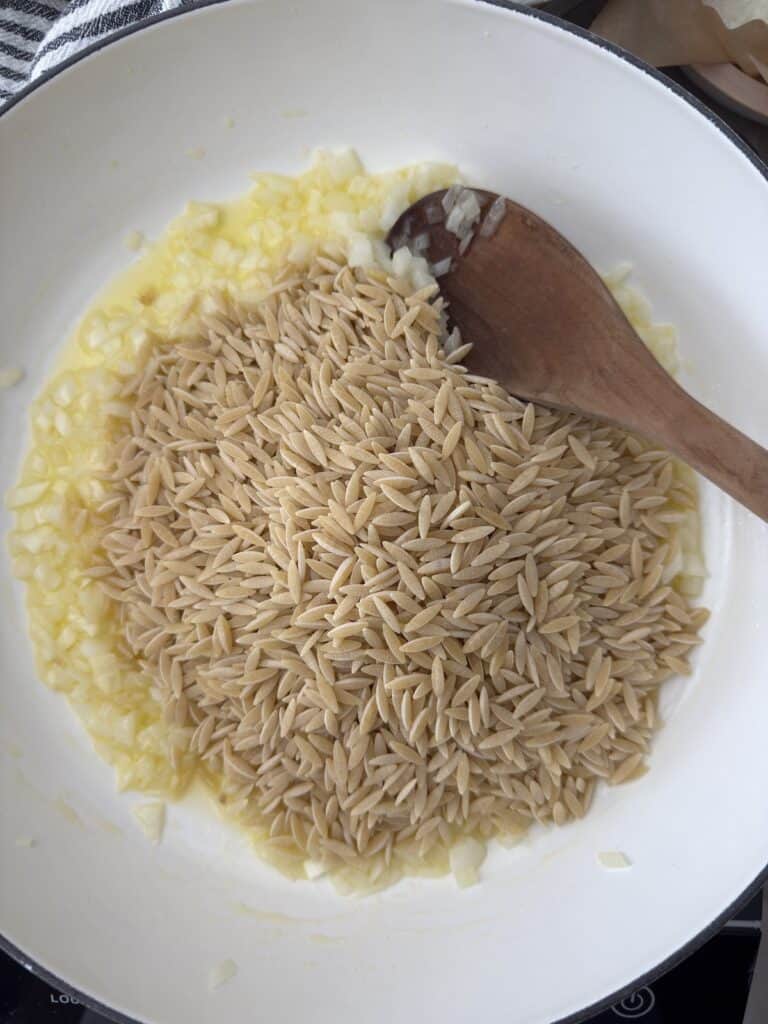 White skillet with the orzo , butter, olive oil and onion with wooden spoon,