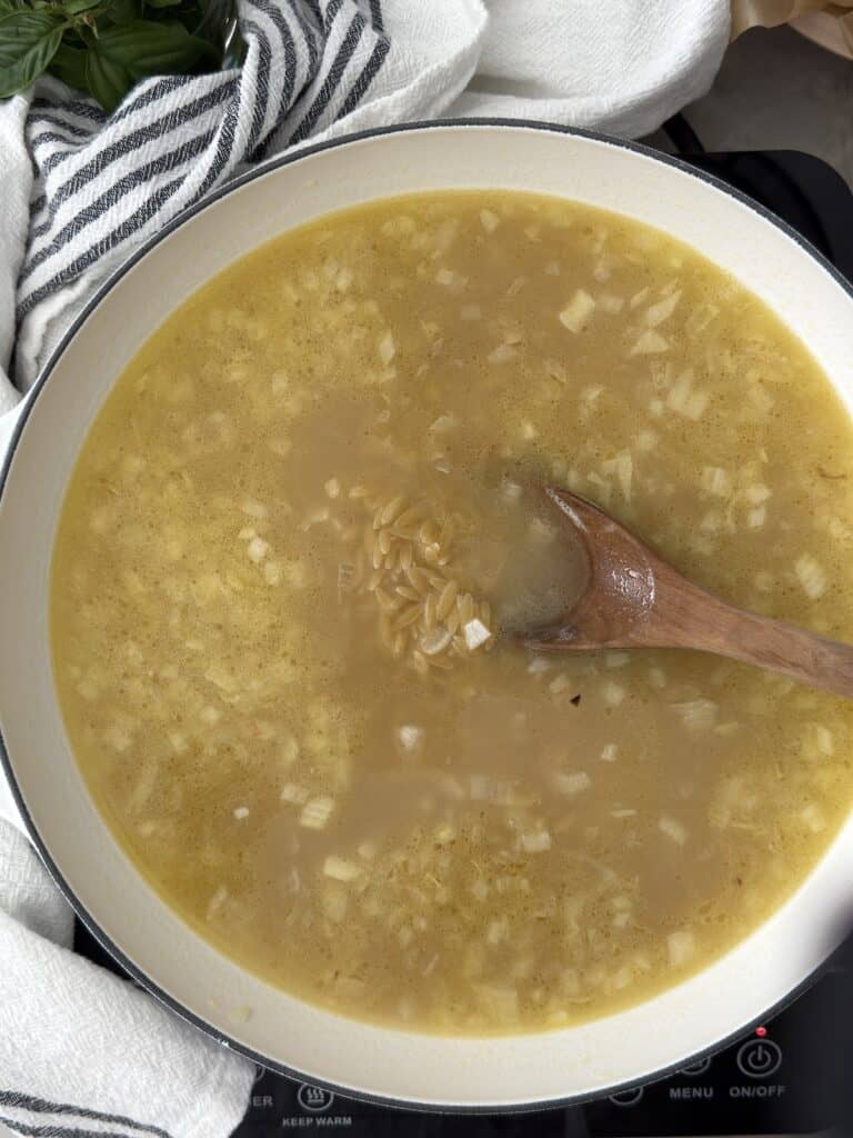 An overhead photo of the orzo in a white skillet just after the chicken broth has been added with wooden spoon to stir.