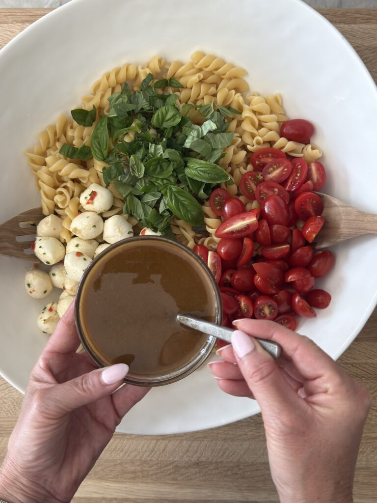 Hands holding a small bowl of vinaigrette dressing above a Caprese pasta salad with rotini, cherry tomatoes, mozzarella pearls, and fresh basil