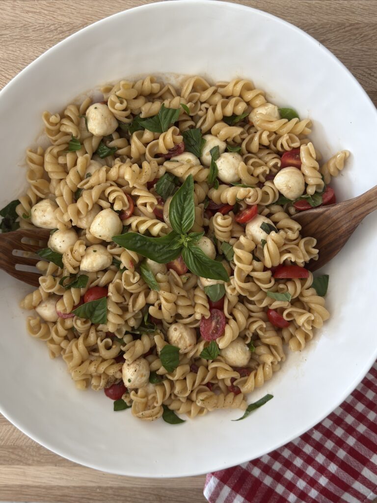 Caprese pasta salad tossed with vinaigrette, mozzarella pearls, cherry tomatoes, and fresh basil in a white bowl with wooden salad utensils.