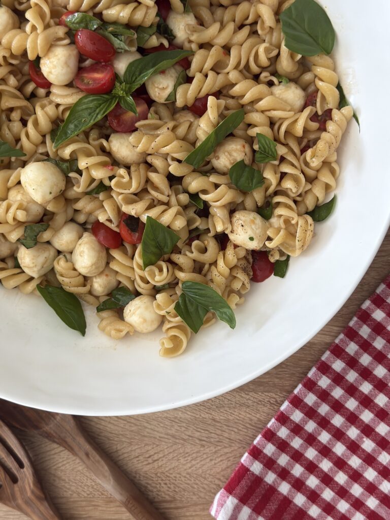 Overhead view of a Caprese pasta salad tossed with rotini, cherry tomatoes, mozzarella pearls, and fresh basil, served in a white bowl on a red gingham napkin.