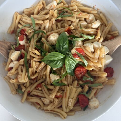 Caprese pasta salad with cherry tomatoes, mozzarella pearls, rotini pasta, and fresh basil leaves tossed with vinaigrette in a white bowl