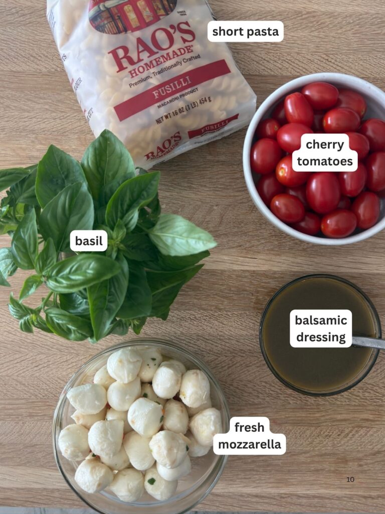 Flat lay of Caprese pasta salad ingredients including fusilli pasta, cherry tomatoes, fresh basil, mozzarella balls, and balsamic dressing on a wooden surface.
