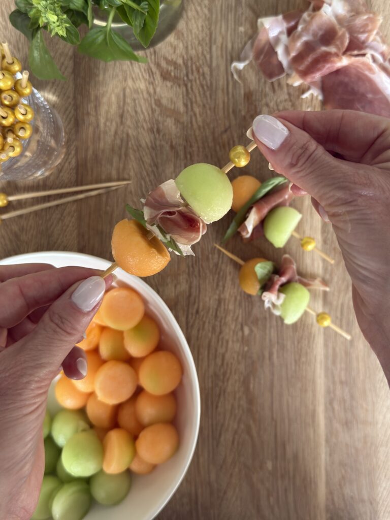 Hands assembling skewers with cantaloupe, honeydew, and prosciutto for a fresh Italian summer appetizer