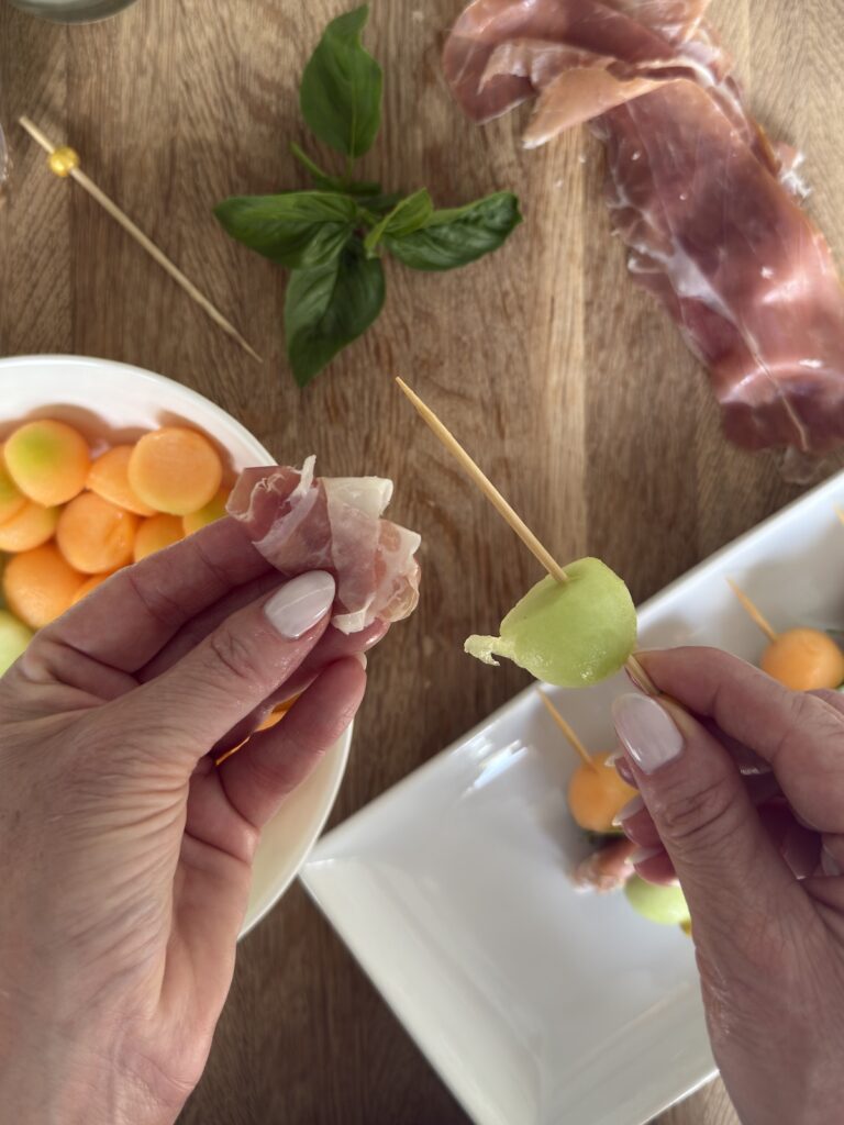 Close-up of hands assembling melon and prosciutto skewers with cantaloupe and honeydew on a wooden board