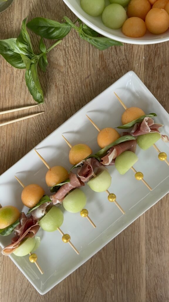 Melon and prosciutto skewers with fresh basil on a white platter, served with a bowl of cantaloupe and honeydew