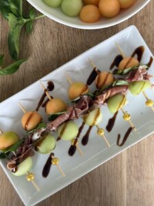Melon and prosciutto skewers with cantaloupe, honeydew, basil, and balsamic glaze on a white serving platter