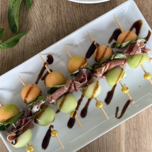 Melon and prosciutto skewers with cantaloupe, honeydew, basil, and balsamic glaze on a white serving platter