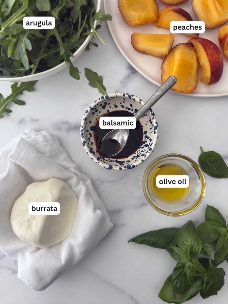 Flat lay of ingredients for grilled peach and burrata salad including fresh arugula, sliced peaches, balsamic vinegar, olive oil, burrata cheese, and basil on a marble surface.