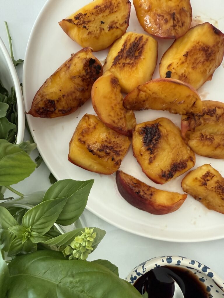 Close-up of freshly grilled peach halves on a white plate, surrounded by fresh basil and salad greens on a marble surface
