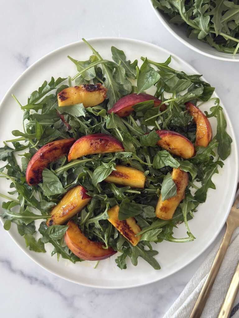 Fresh arugula salad topped with grilled peaches on a white plate, styled with gold flatware and a neutral napkin on a marble surface.
