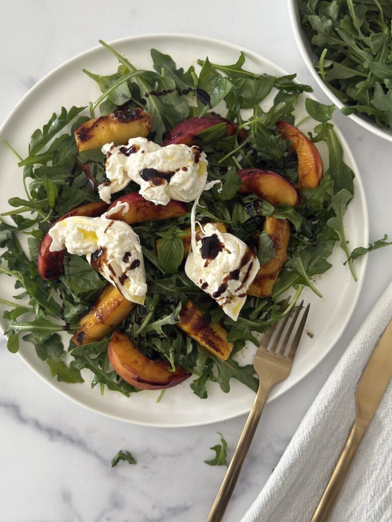Grilled peach and burrata salad with arugula on a white plate, topped with balsamic glaze and served with a gold fork on a marble background.