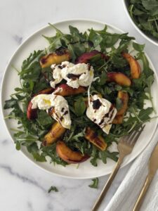 Grilled peach and burrata salad with arugula on a white plate, topped with balsamic glaze and served with a gold fork on a marble background.