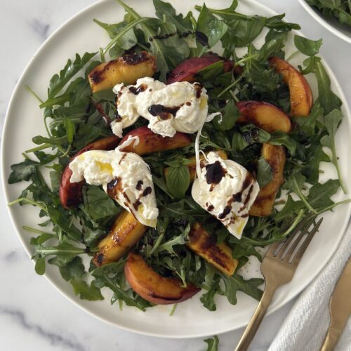 Grilled peach and burrata salad with arugula on a white plate, topped with balsamic glaze and served with a gold fork on a marble background.