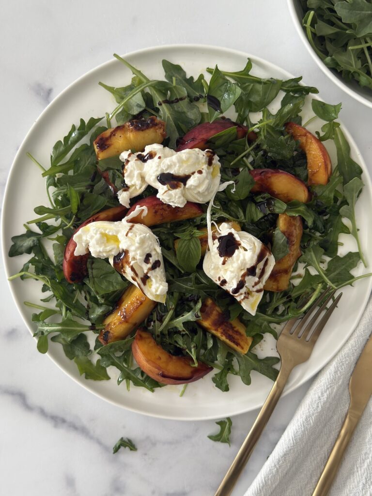 Grilled peach and burrata salad with arugula on a white plate, topped with balsamic glaze and served with a gold fork on a marble background.