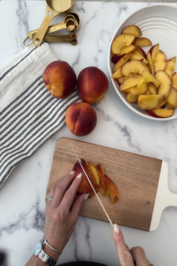 Hands slicing fresh peaches on cutting board with whole peaches and bowl of sliced fruit nearby — prep step for homemade peach crostata recipe.