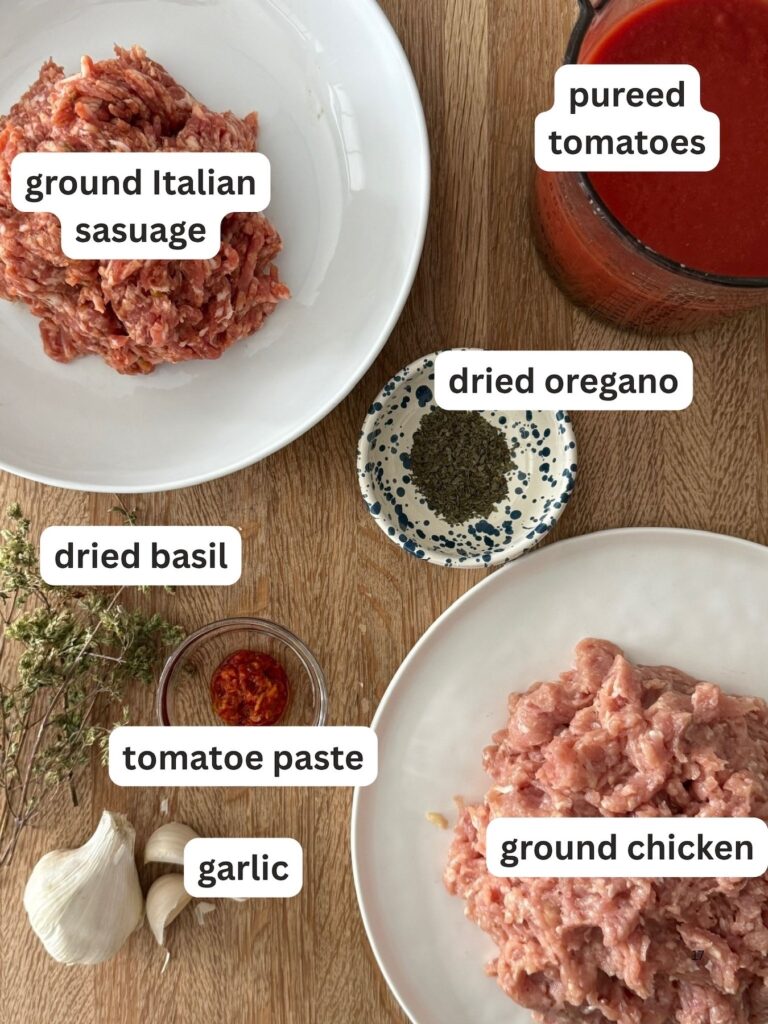 Flat lay of Italian meat sauce ingredients including ground Italian sausage, ground chicken, pureed tomatoes, tomato paste, garlic, dried oregano, and dried basil on a wooden board