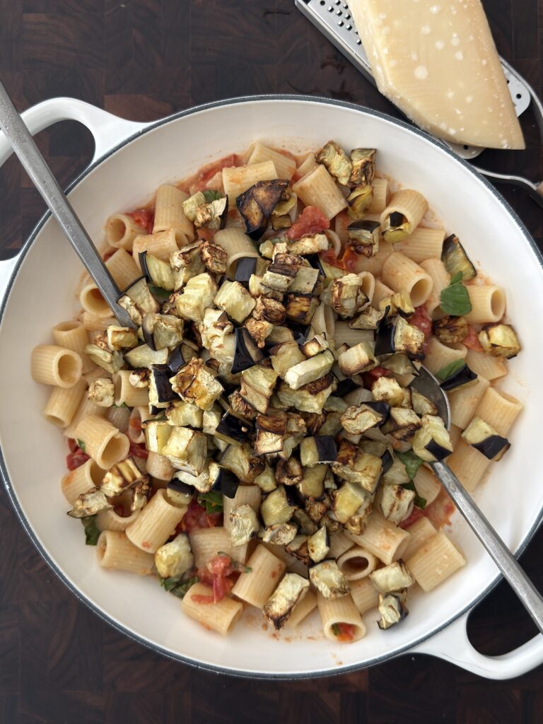 Creamy eggplant pasta in a white skillet topped with roasted eggplant, cherry tomato sauce, fresh basil, and Parmesan cheese before stirring.