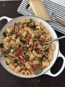 Creamy eggplant pasta in a white skillet topped with roasted eggplant, cherry tomato sauce, fresh basil, and Parmesan cheese before stirring.