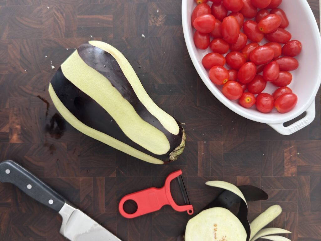 Raw eggplant being peeled on a cutting board with a knife and vegetable peeler, next to a white bowl of cherry tomatoes, for creamy eggplant pasta.