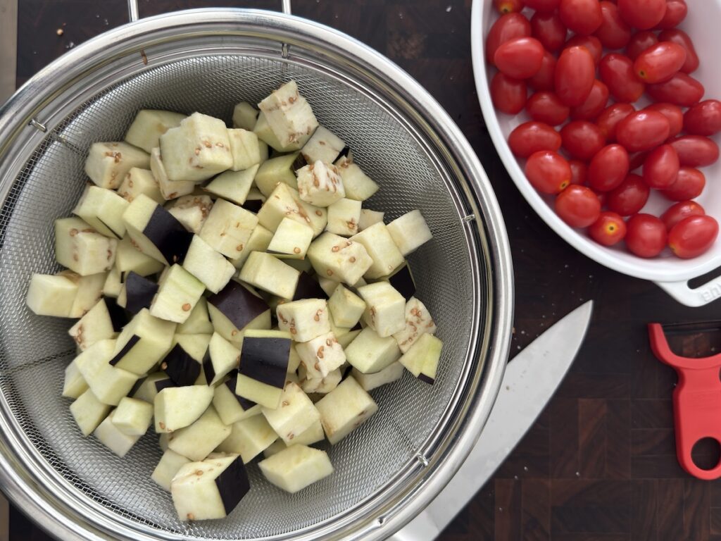 Cubed eggplant draining in a colander next to a bowl of cherry tomatoes, prepped for creamy eggplant pasta.