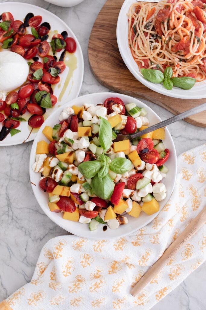 Bowl of summer peach Caprese salad with cherry tomatoes, mozzarella pearls, cucumber slices, and fresh basil leaves on a linen background