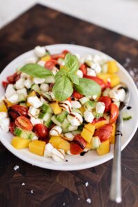 Overhead shot of peach caprese salad with cherry tomatoes, fresh mozzarella pearls, cucumber, and basil, drizzled with balsamic glaze in a white bowl
