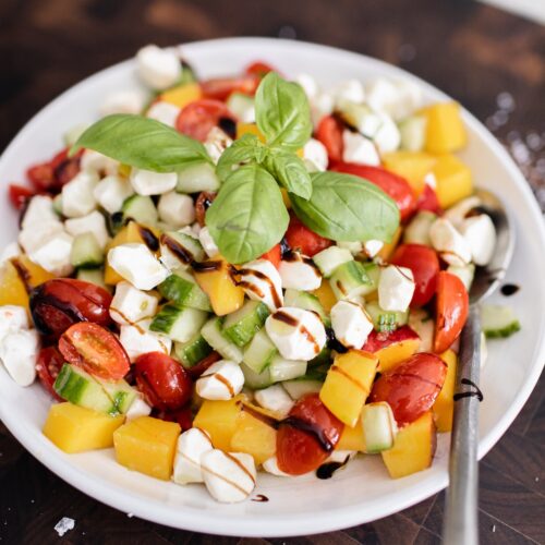 Overhead shot of peach caprese salad with cherry tomatoes, fresh mozzarella pearls, cucumber, and basil, drizzled with balsamic glaze in a white bowl