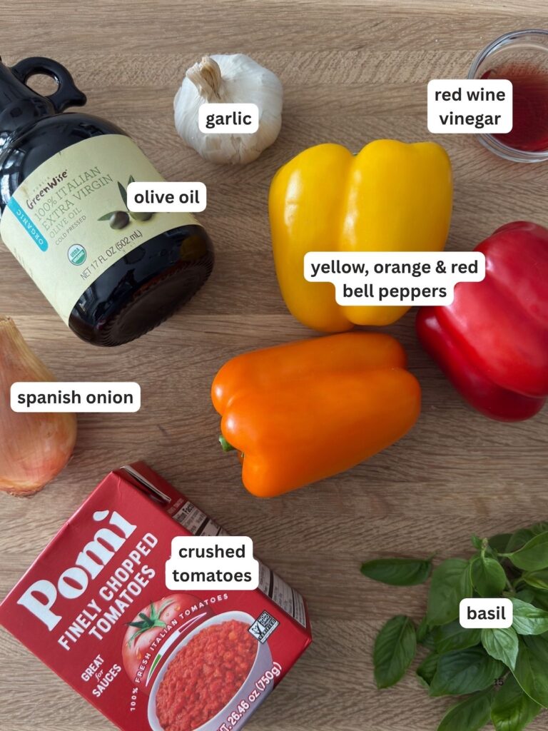 Ingredients for Italian pepperonata – red, yellow, and orange bell peppers, onion, garlic, basil, crushed tomatoes, olive oil, and red wine vinegar on a wooden board